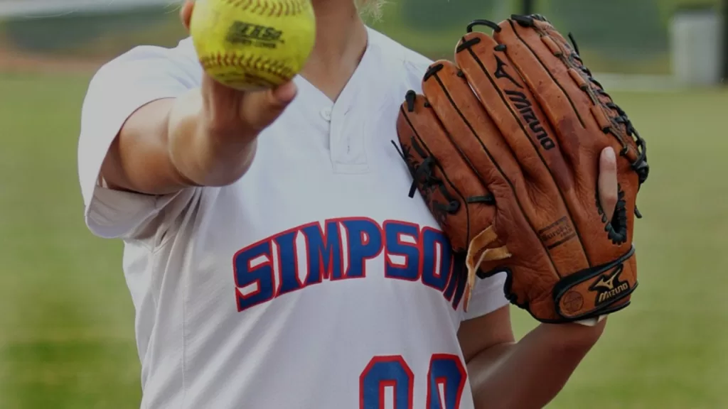 Woman in baseball gear holding up baseball to camera